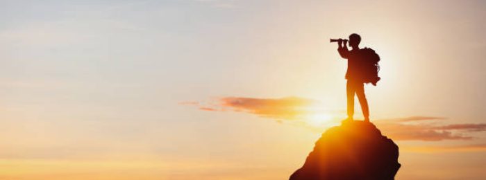 istockphoto-1383796215-612×612 vision for success ideas. businessman's perspective for future planning. Silhouette of man holding binoculars on mountain peak against bright sunlight sky background.